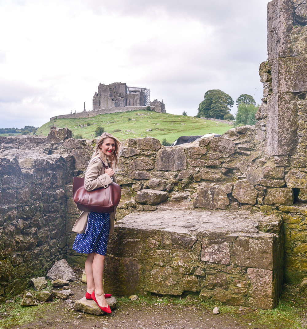 Jenna Colgrove in a navy polka dotted dress and red flats with the Rock of Cashel in Ireland. 
