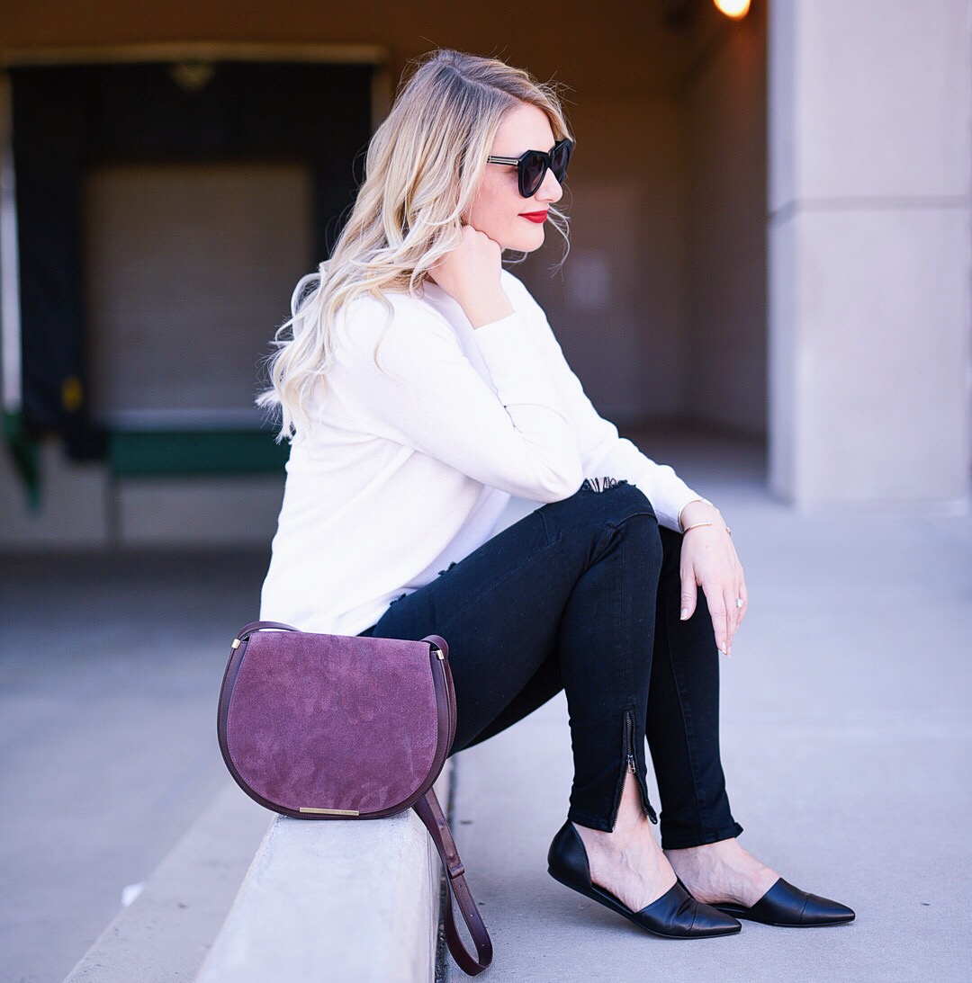 Jenna Colgrove wearing a white cashmere sweater, black zip skinny jeans, and a Cuyana Saddle Bag in Oxblood. 