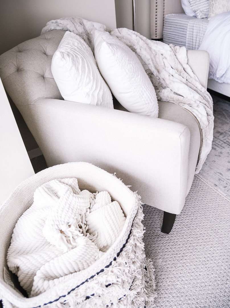 An Anthropologie hamper, a throw blanket, and white frayed pillows. 